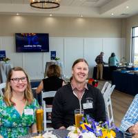 Group of three sitting at table at event, pausing meal to smile at camera